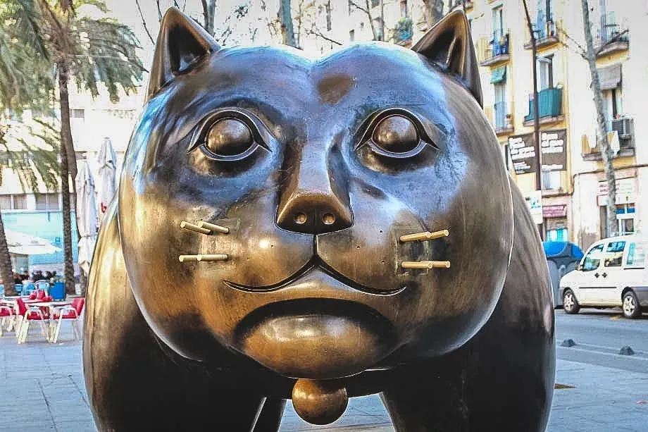 Large bronze sculpture of a fat cat with round eyes and brass whiskers on a palm-lined street in Barcelona.