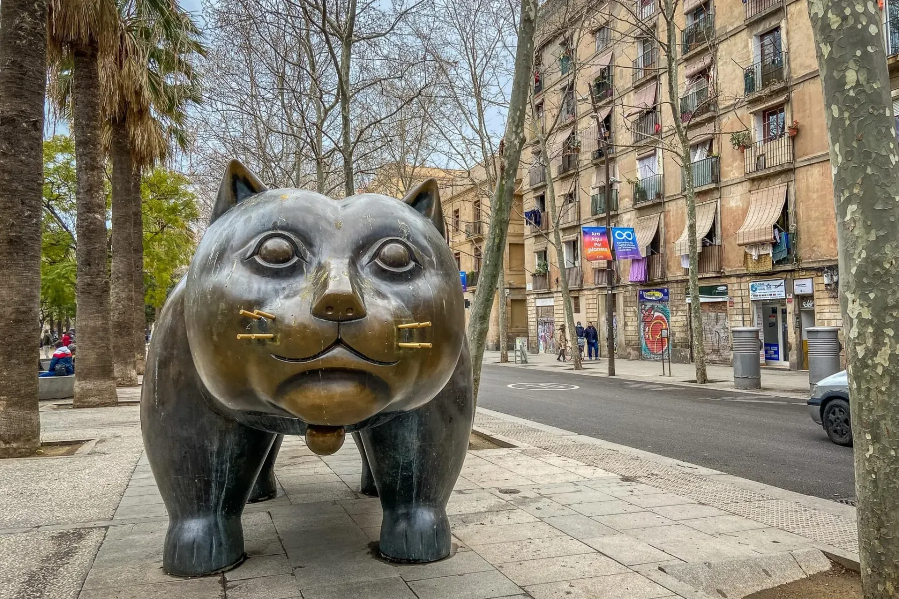 Large bronze sculpture of a plump cat with small ears, on a city sidewalk next to a street with buildings and trees.