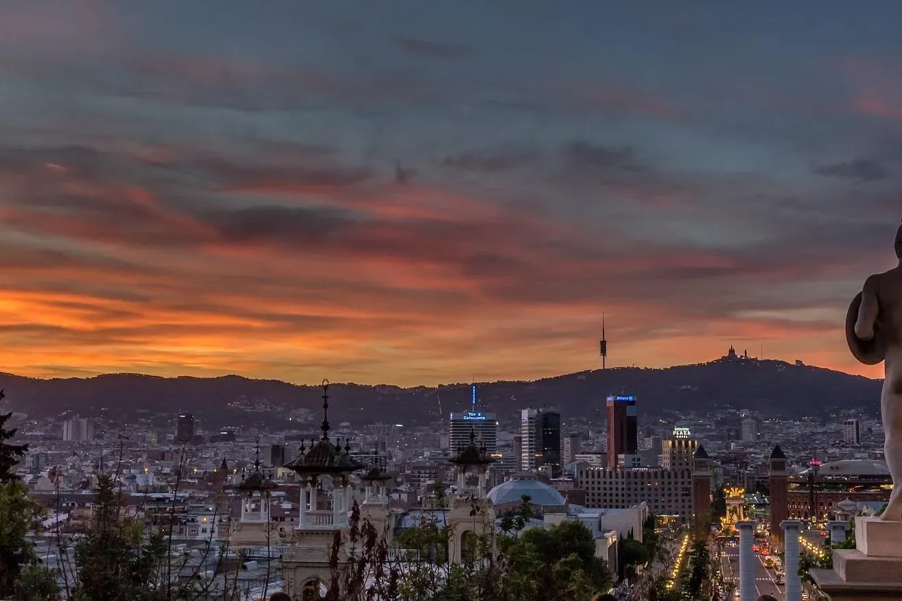 View of Barcelona city skyline at sunset with a statue in the foreground and Tibidabo mountain in the background.