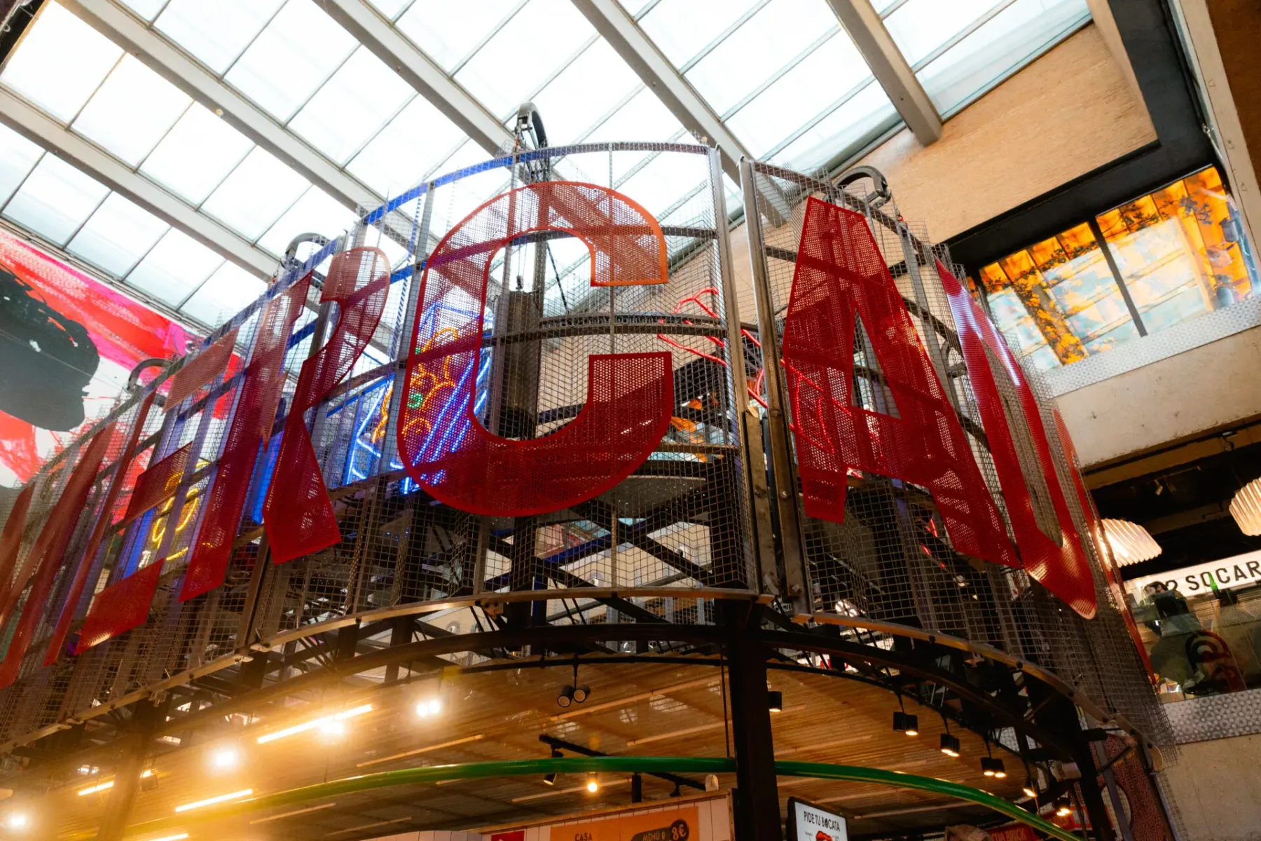 Large red perforated metal letters spelling "ARCELÓ" on a mesh structure with blue neon lights, under a glass roof.
