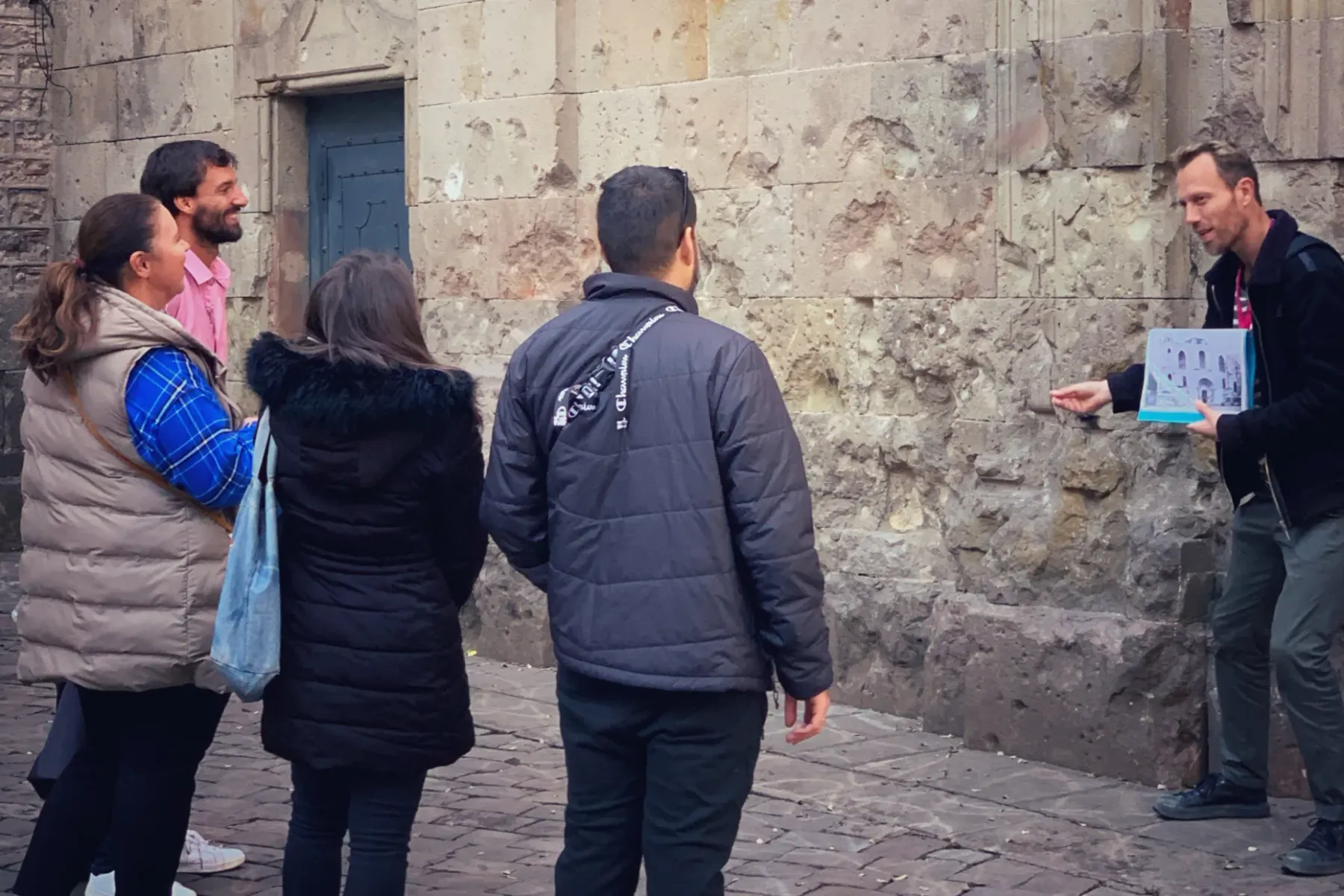 A male guide holding a brochure points at an old stone wall, speaking to three tourists.