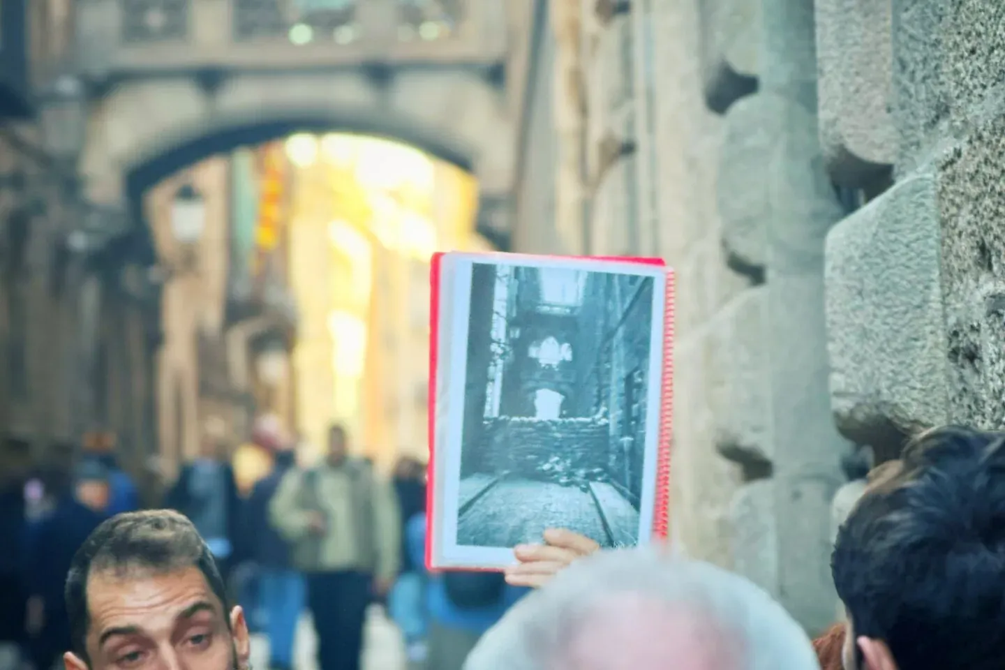 Male tour guide showing an old black-and-white photo of a street to tourists, with historic architecture and an arched bri...
