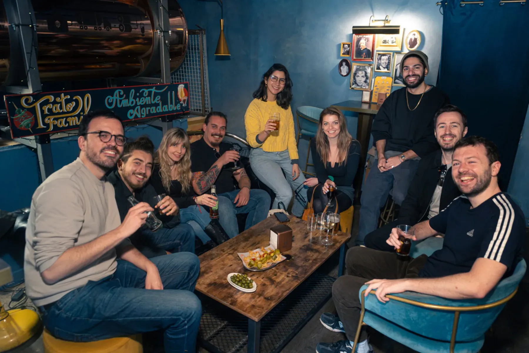 Group of diverse friends smiling in a dimly lit bar, holding drinks around a wooden table with snacks. A large copper tank...