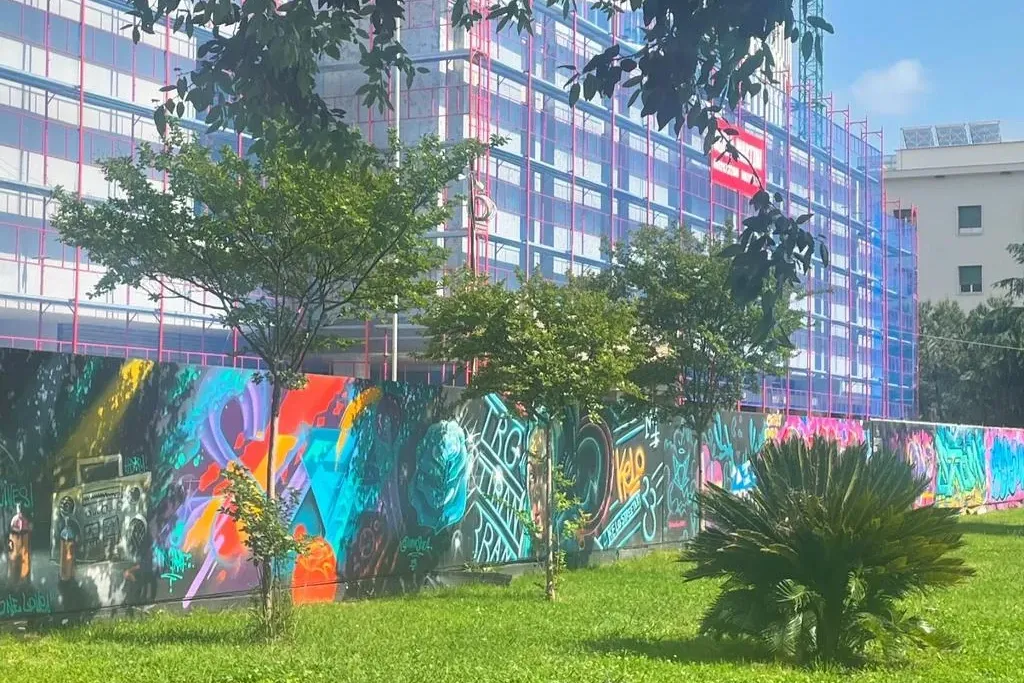 Colorful street art mural on a wall with green grass in foreground, a building under construction, and trees.
