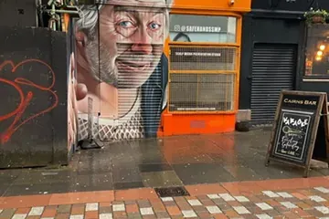 Large mural of an old man's face with grey hair on a building wall, partially covering a metal shutter. Wet brick paving i...