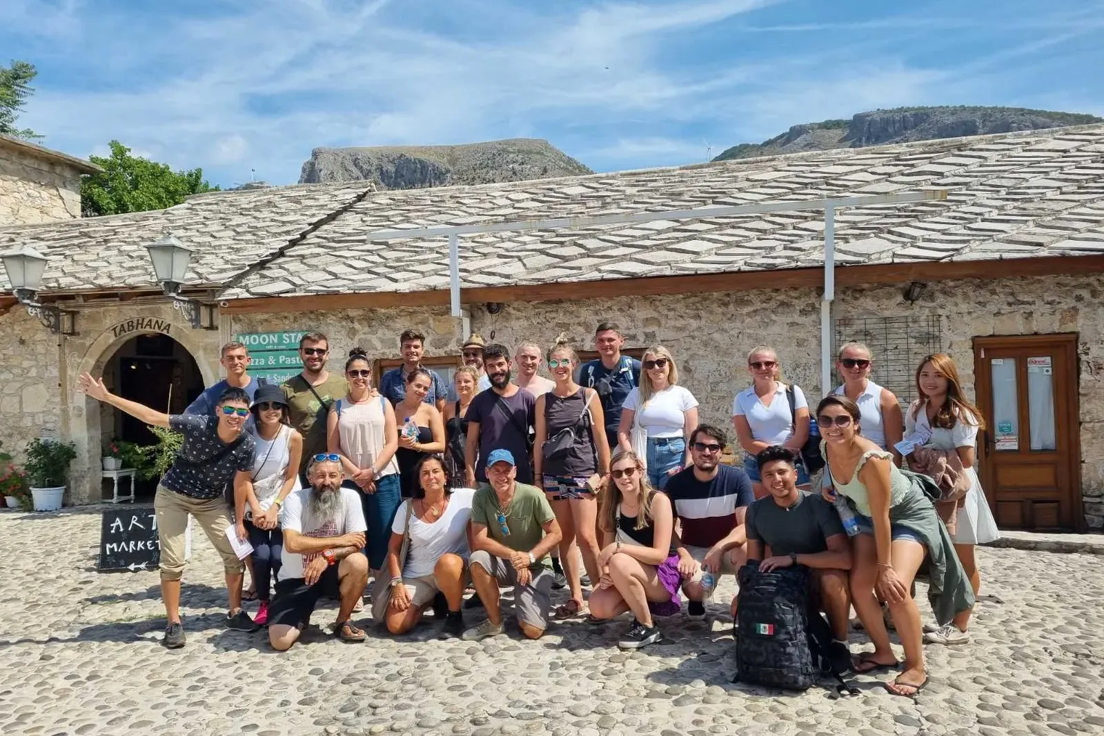 A diverse group of tourists poses smiling on a cobblestone street in front of a traditional stone building with a slab roo...