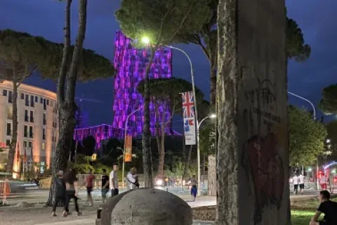 Concrete bunker and a segment of the Berlin Wall with graffiti in a public square at dusk in Tirana. People are walking in...