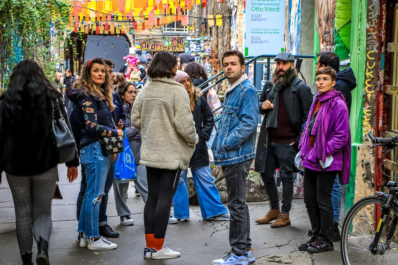 Group of people in a narrow, art-covered alley with graffiti walls and colorful banners overhead. A museum sign is visible.