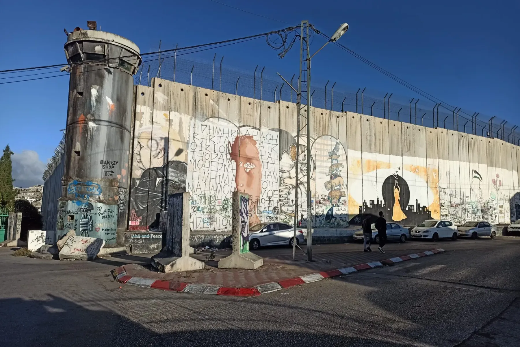 Concrete separation wall with watchtower covered in graffiti and murals, cars parked, under a clear blue sky in Bethlehem.