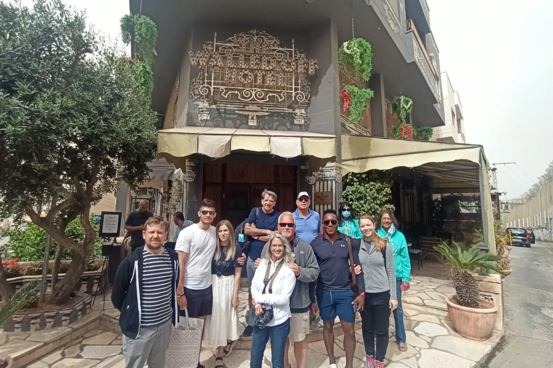Group of ten tourists posing in front of The Walled Off Hotel in Bethlehem, with an olive tree and potted plants.