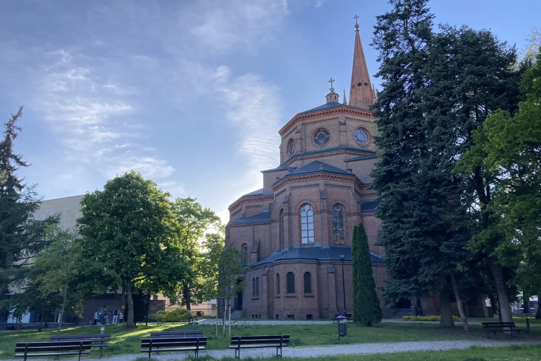 Large brick church with a tall steeple, seen from a green park with trees and benches under a partly cloudy sky.