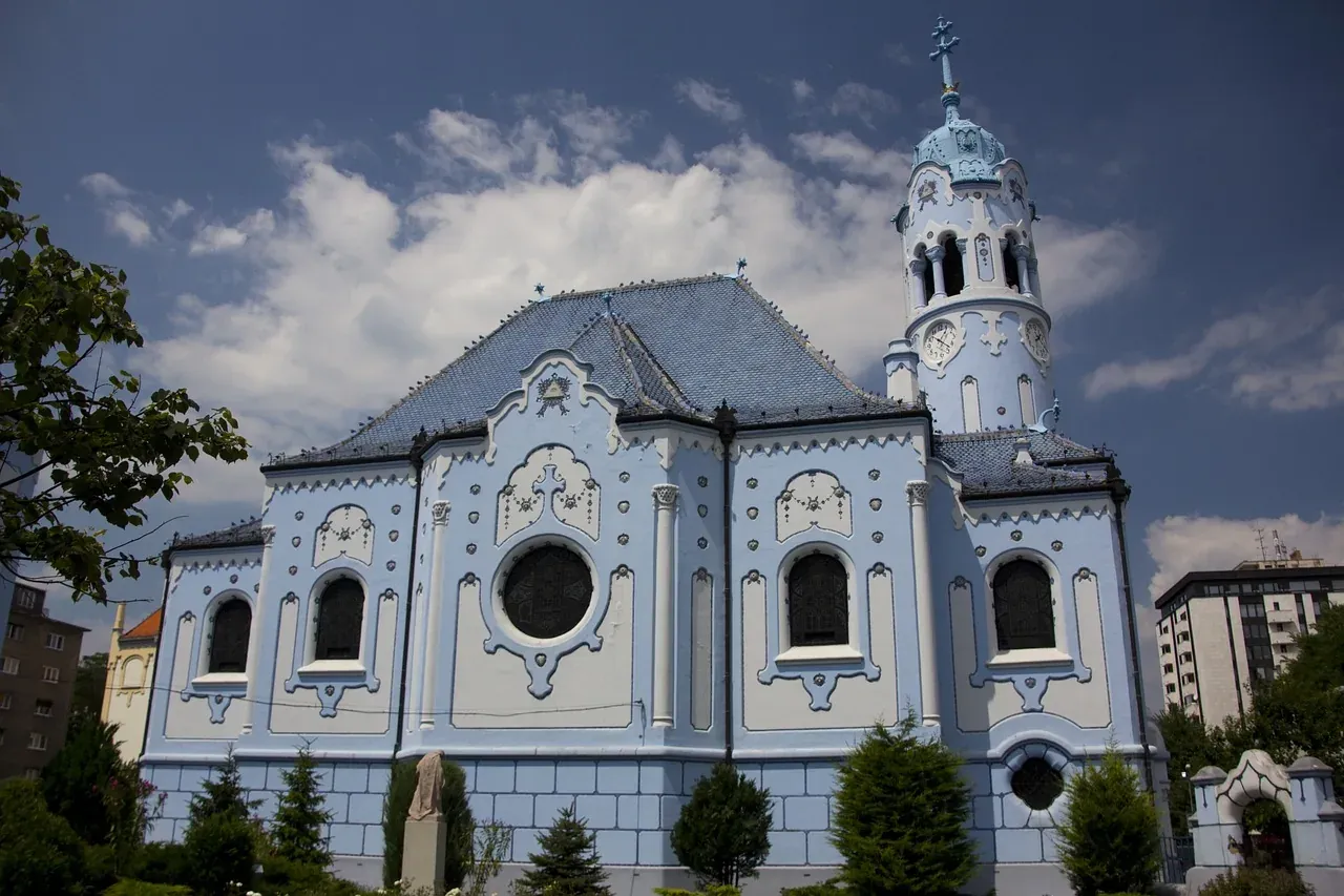 The light blue Church of St. Elisabeth, known as the Blue Church, with a distinctive blue roof and tower under a cloudy sky.