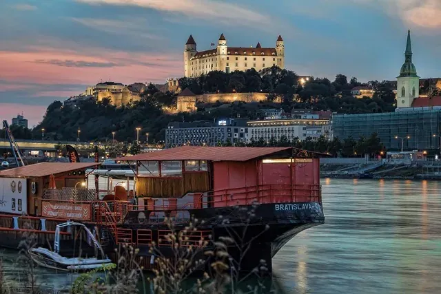 Bratislava Castle and St. Martin's Cathedral illuminated at dusk. A floating bar is on the Danube River in the foreground.