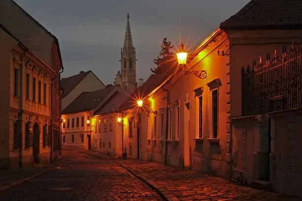 Cobblestone street lined with old buildings at night, illuminated by warm streetlights, with a church spire visible in the...