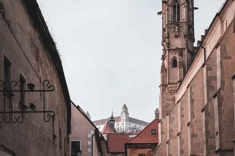 Cobblestone street leading towards St. Martin's Cathedral tower and Bratislava Castle on a cloudy day.