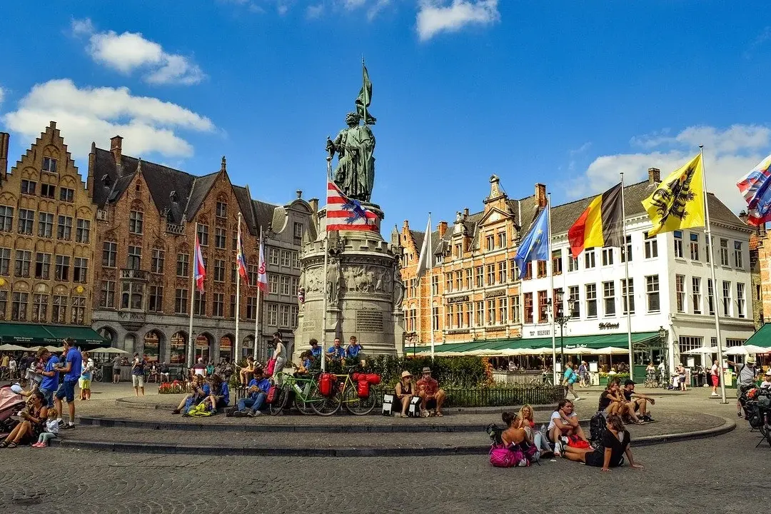 Historic Markt square in Bruges with the Hendrik Conscience statue, gabled buildings, national flags, and many visitors.