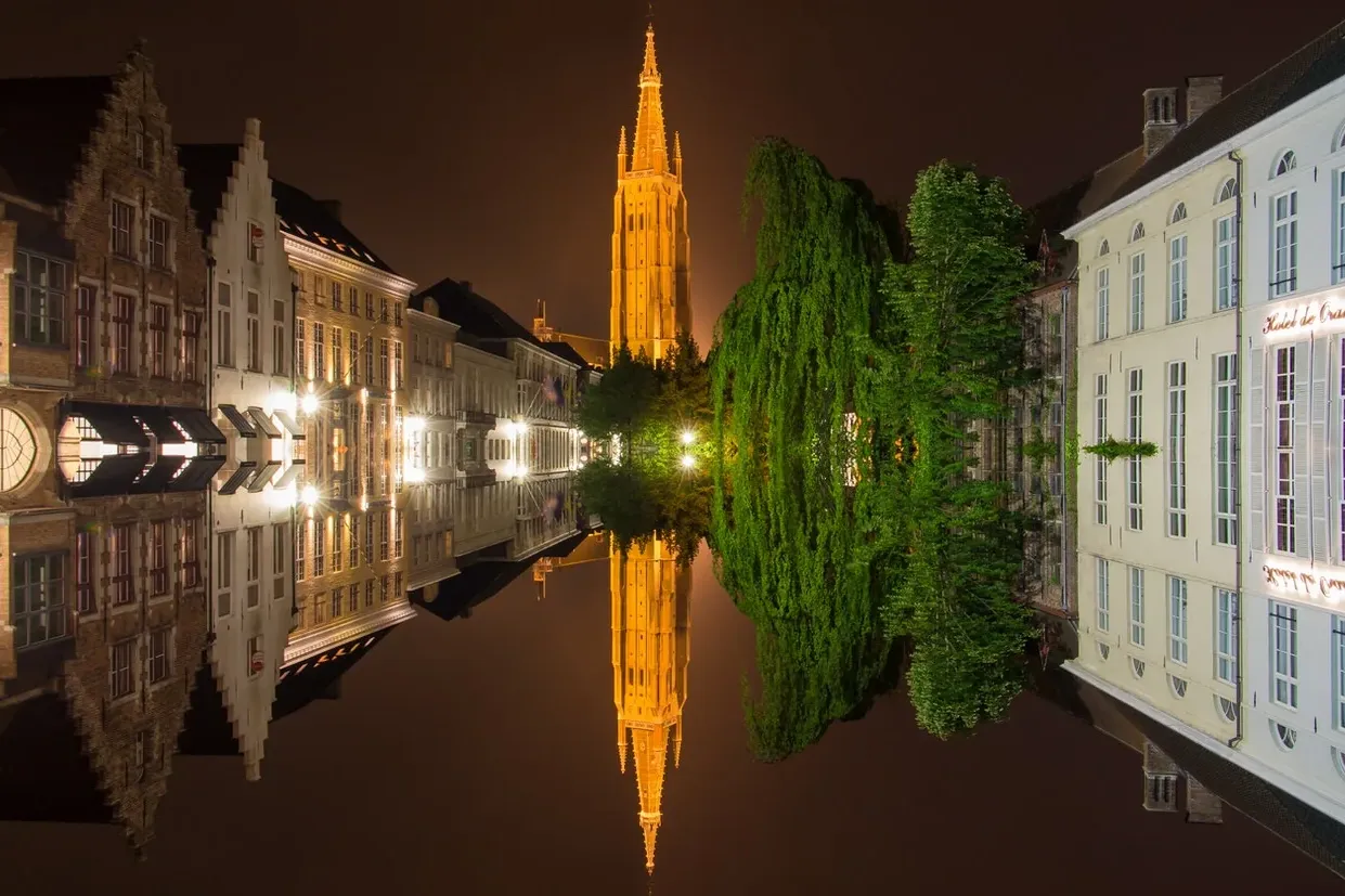 Illuminated historic buildings and the Belfry tower reflected in a calm Bruges canal at night.