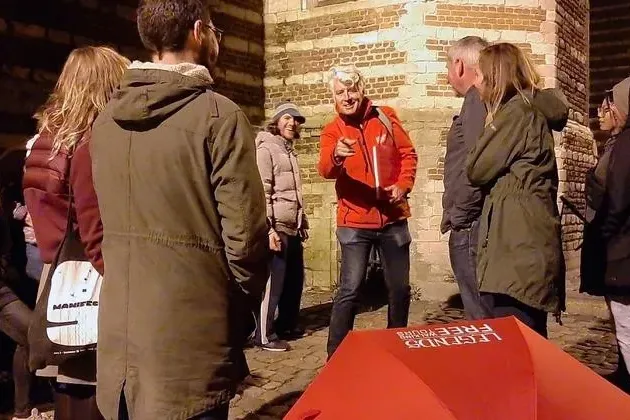 A tour guide in a red jacket pointing, addressing a group of tourists at night. A red umbrella is visible, with an old bri...