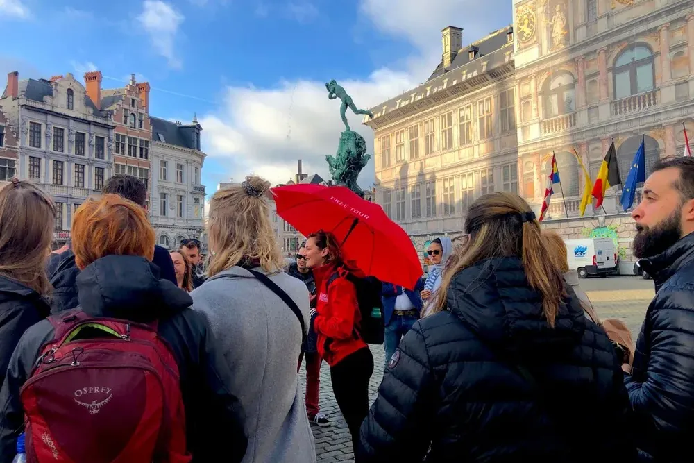 A group of people listening to a tour guide with a red umbrella in a historic square with the Brabo Fountain and City Hall...