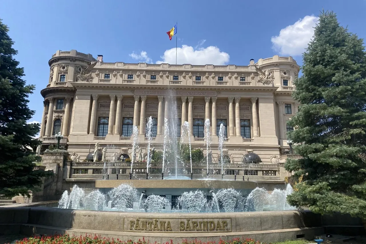 Neo-classical building with columns, round towers, and a Romanian flag, in front of a multi-tiered fountain with water jets.