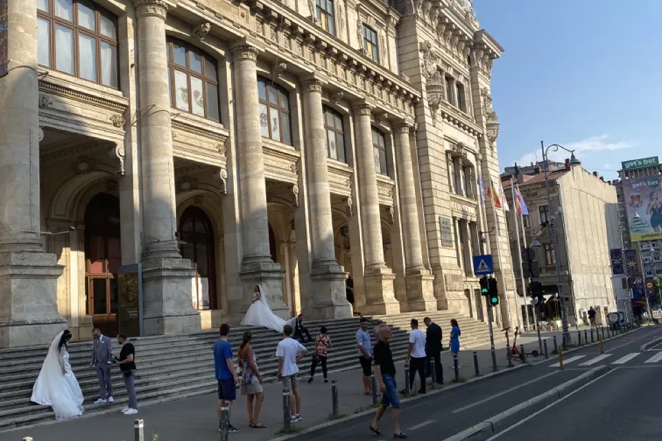 Grand stone building with columns, steps, and a dome. Two wedding couples and pedestrians are on the sidewalk and steps. S...