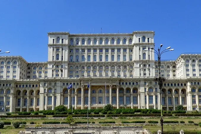 Massive, ornate white stone building with many windows and columns under a clear blue sky, seen from a distance with green...