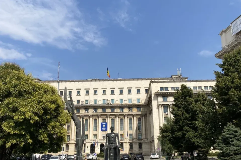Large building with a Romanian flag, two dark modern sculptures in a paved square, and green trees under a blue sky.