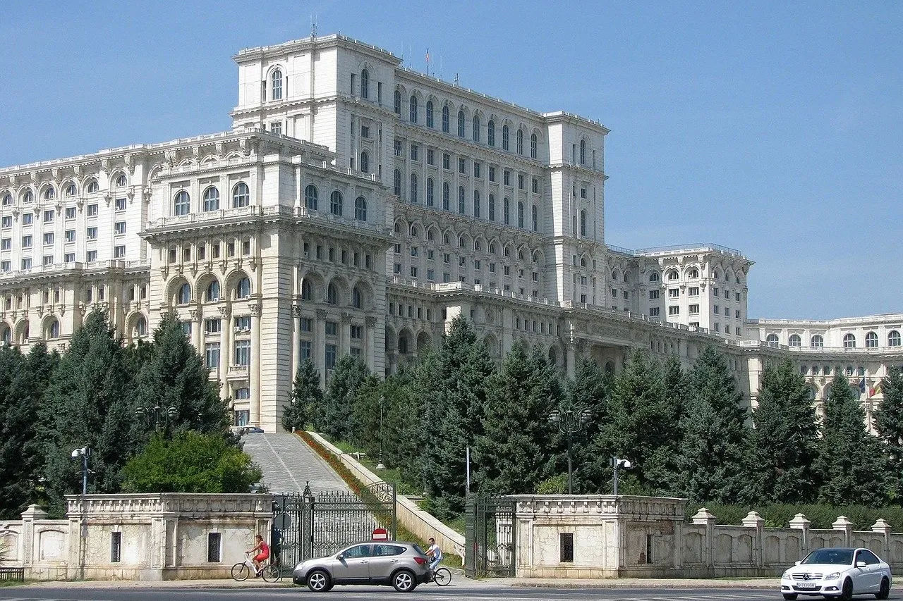 Massive white Palace of the Parliament in Bucharest, partially obscured by green trees, with a street and two cars in the ...