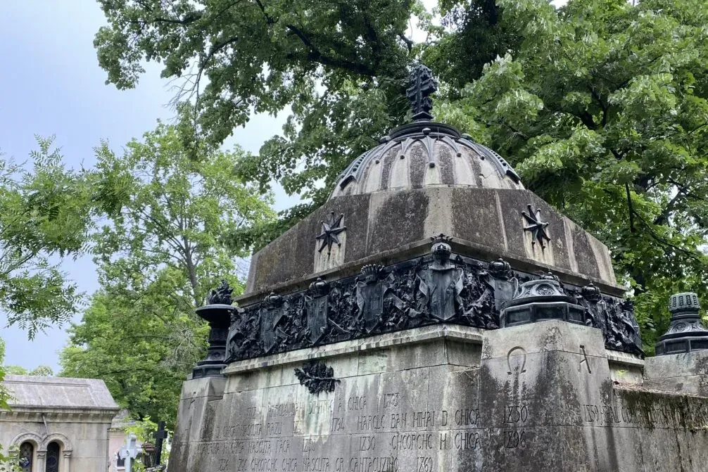 Ornate stone mausoleum with a dome and cross finial, detailed relief sculptures, and inscriptions, surrounded by green tre...