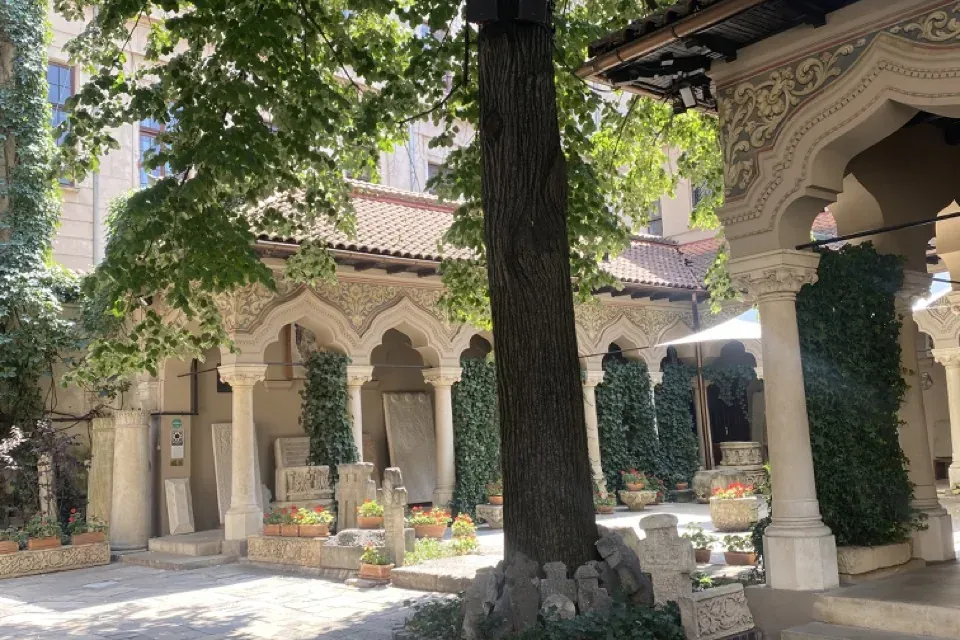 Paved courtyard of a monastery with a large tree, arched colonnades, and various old stone sculptures displayed around.