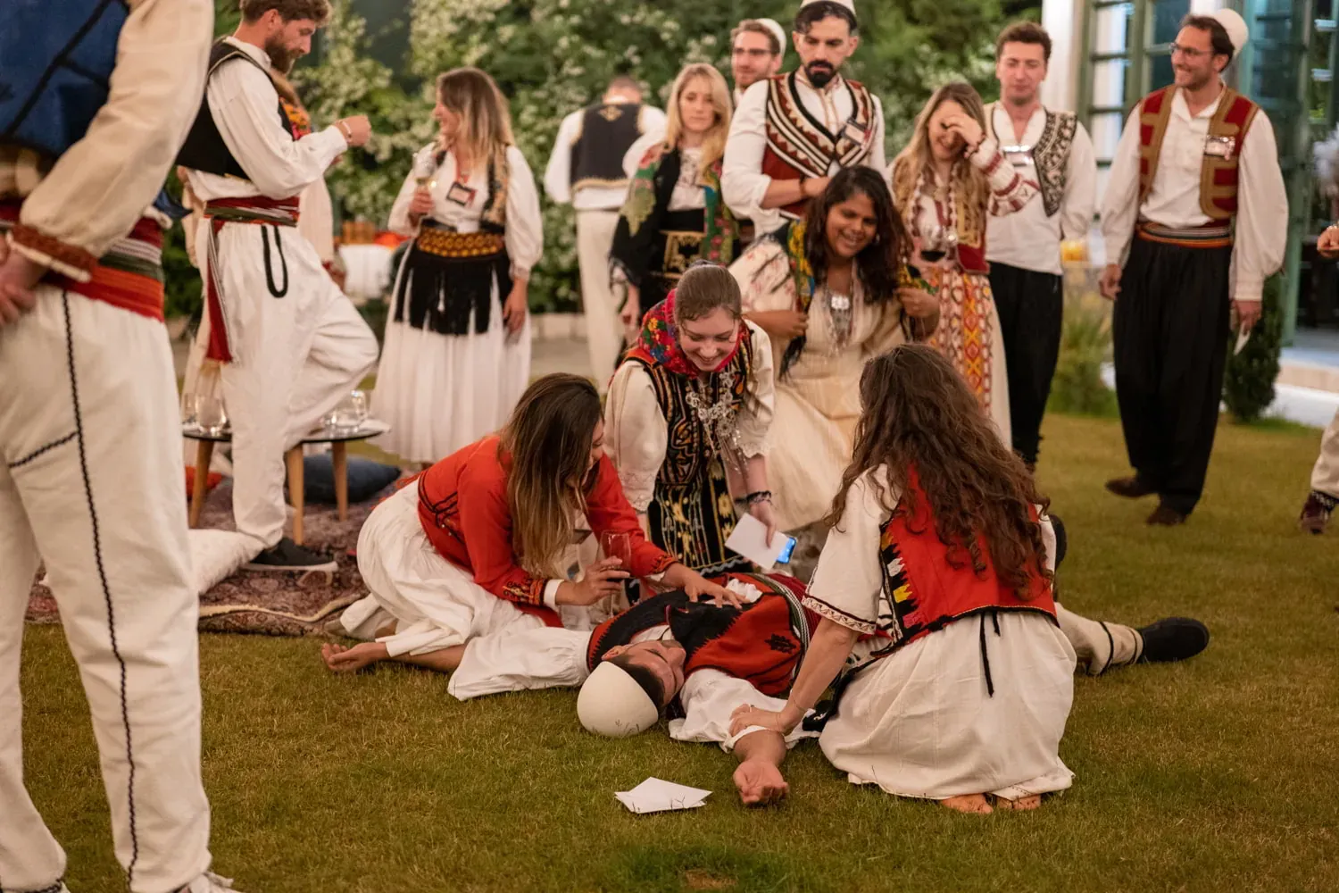 People in traditional Albanian attire participate in an outdoor cultural performance on grass. Two women kneel beside a ma...