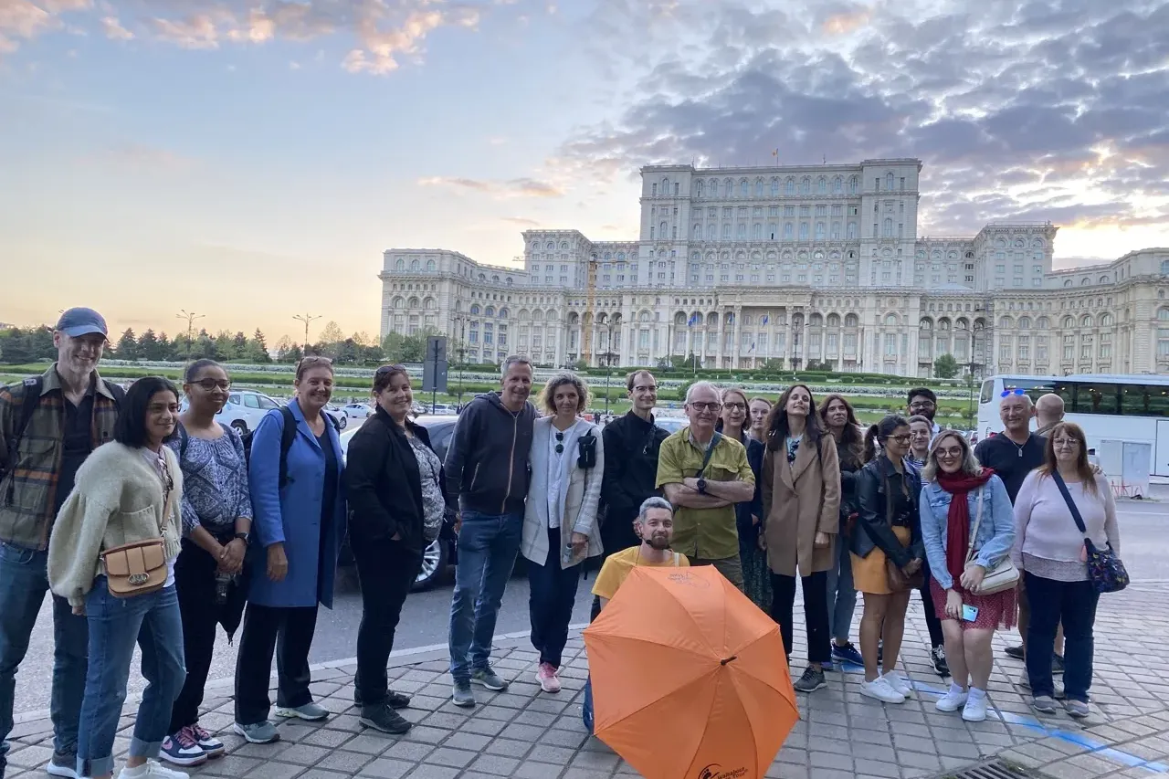 A group of diverse people stands outdoors in front of the massive Palace of the Parliament in Bucharest under a cloudy sky.