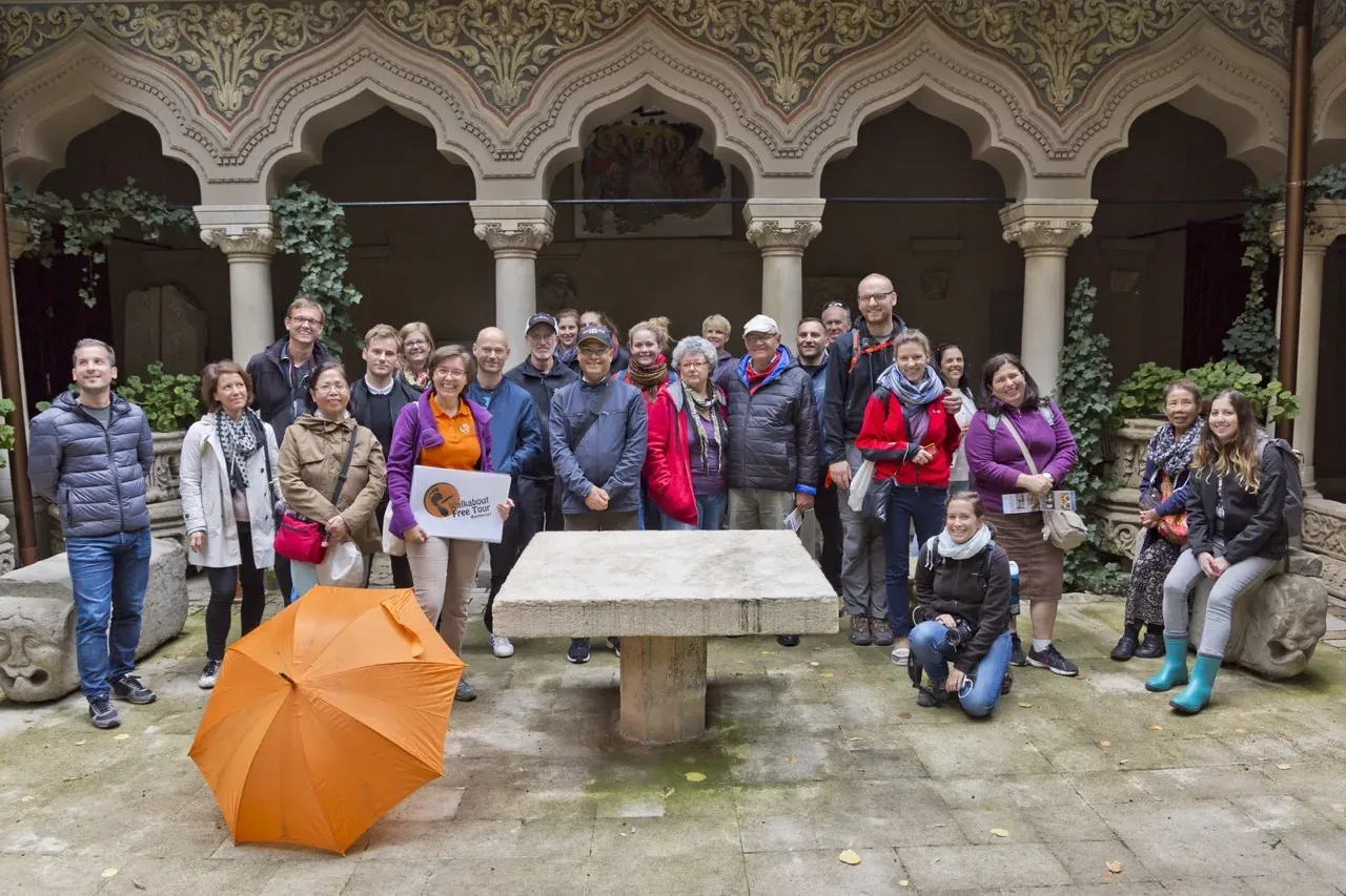 A group of tourists with a guide in the ornate arched courtyard of Stavropoleos Monastery in Bucharest, with an orange umb...