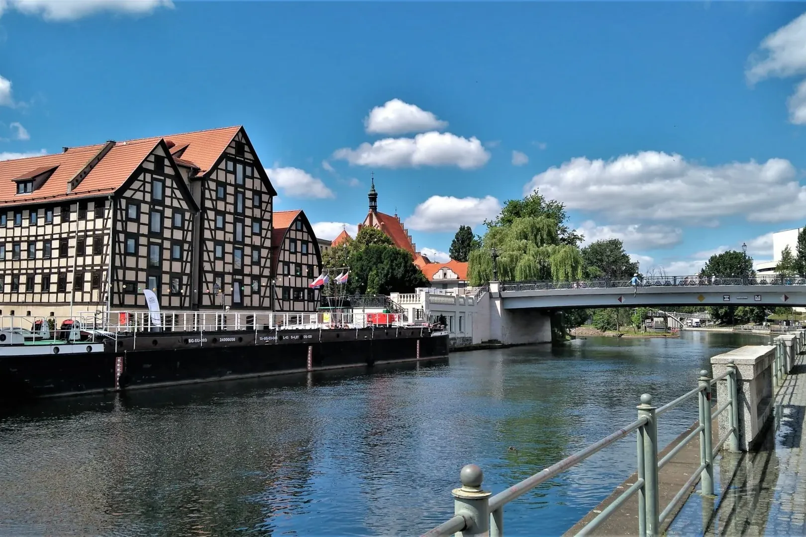 Half-timbered buildings with red roofs on the riverbank, a long barge, a concrete bridge, and a pedestrian path under a bl...