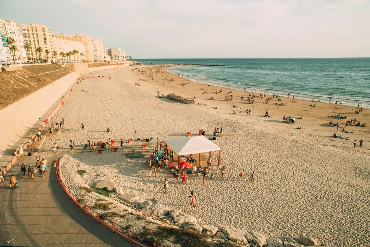 Wide sandy beach in Cadiz with many people, a white tent, blue sea, and coastal buildings under a clear sky.