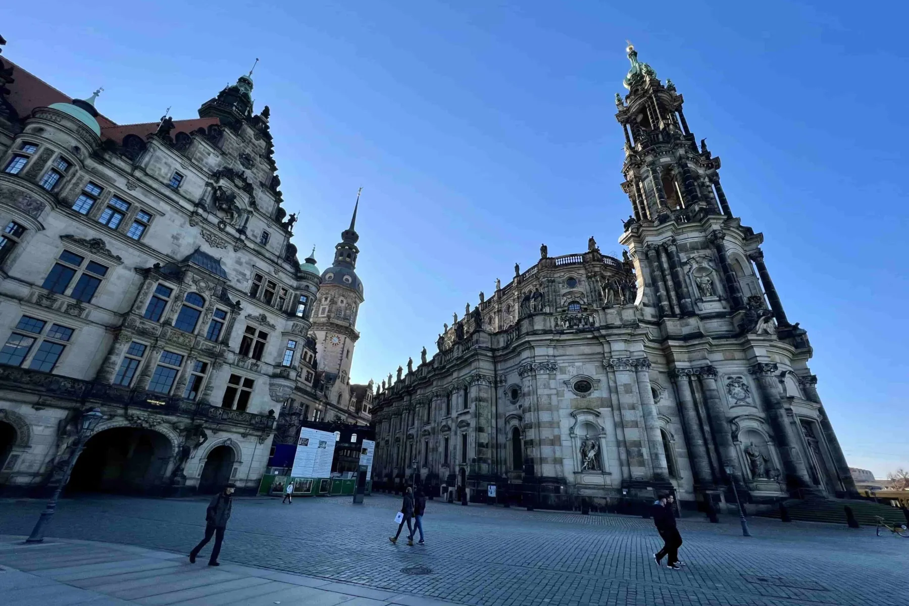 Historic Dresden Royal Palace and Hofkirche under a clear blue sky, with people walking on the cobblestone square.