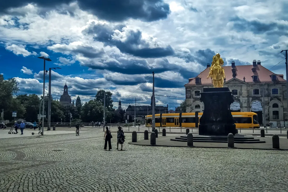 Golden Rider equestrian statue in Dresden, cobblestone square, a yellow tram, and distant city domes under a cloudy sky.