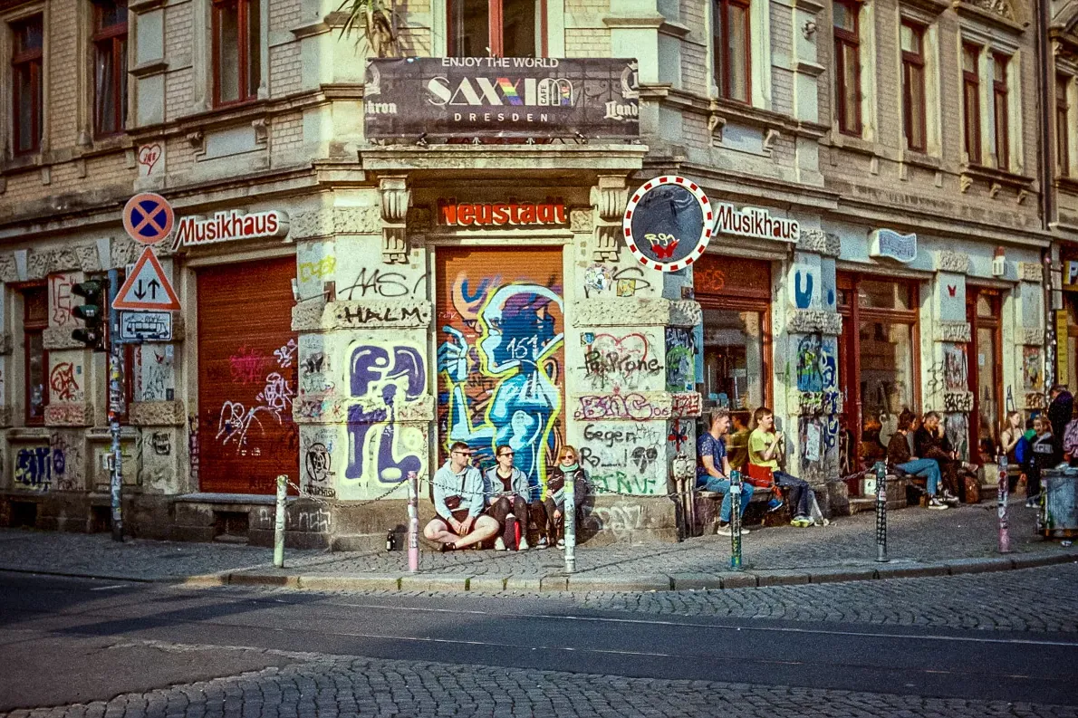 Building corner in Dresden's Neustadt district covered in colorful street art and graffiti. People are sitting on the side...