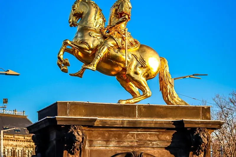 Golden equestrian statue of a man on a rearing horse atop a dark, inscribed pedestal against a blue sky.