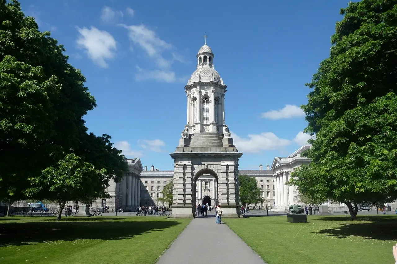 The white stone Campanile of Trinity College Dublin stands amidst green lawns and historic buildings under a bright blue sky.