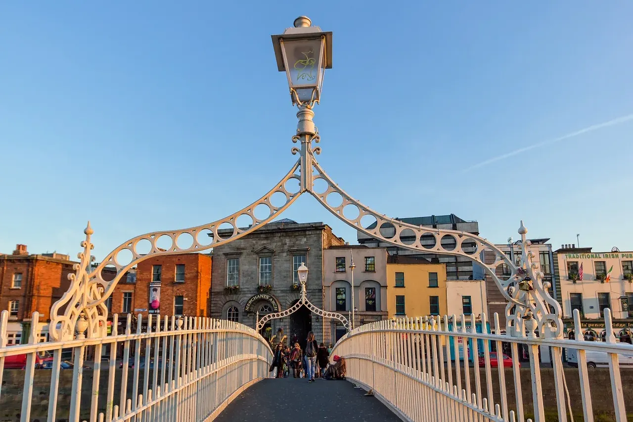White cast-iron Ha'penny Bridge in Dublin at sunset, with pedestrians crossing and historic buildings on the far bank of t...