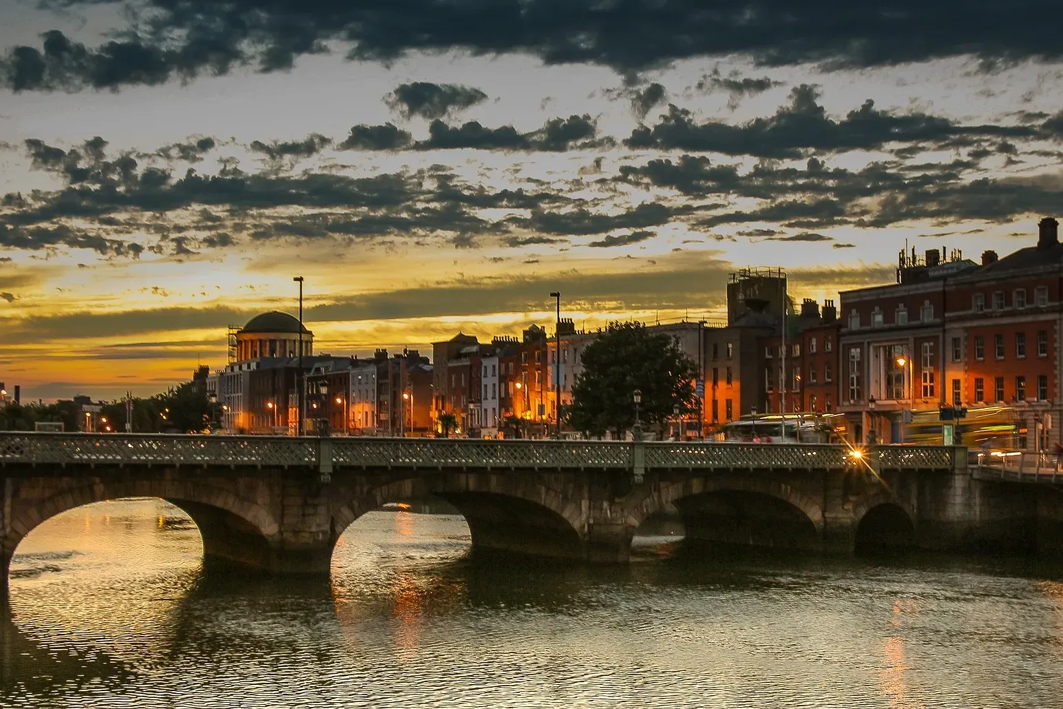 Stone arch bridge over a river in Dublin at sunset, with city buildings lit up on the far bank and a cloudy sky.