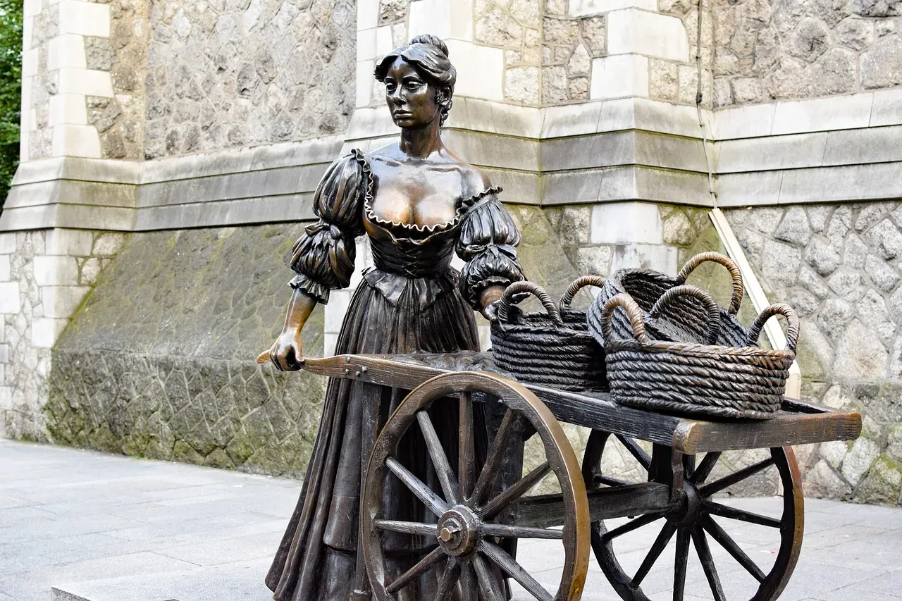 Bronze statue of a woman in historical clothing pushing a two-wheeled cart filled with three woven baskets. Stone building...