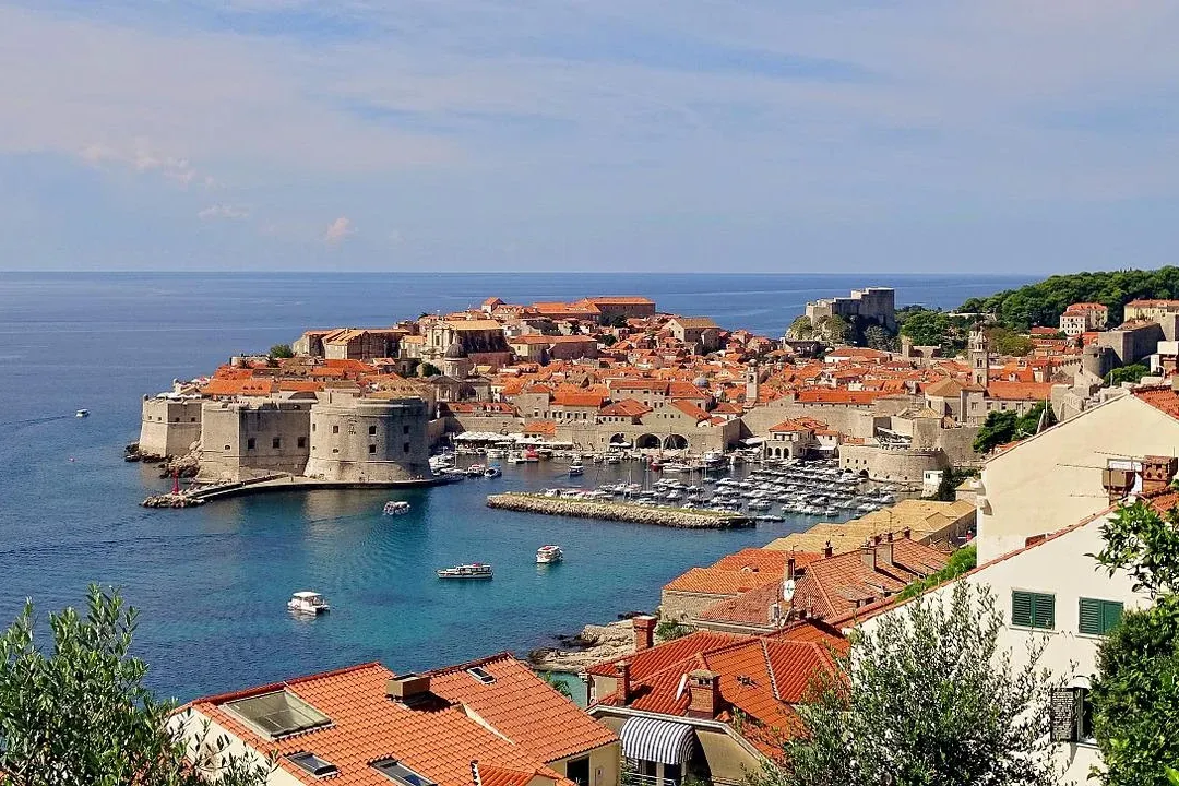 Elevated view of Dubrovnik's Old Town, featuring red-tiled roofs, medieval city walls, a harbor with boats, and the Adriat...