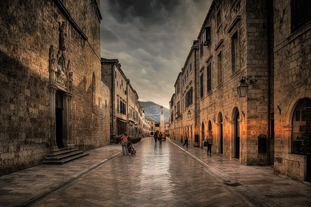 Wet cobblestone street in Dubrovnik Old Town lined with historical stone buildings. A bell tower is visible in the distanc...