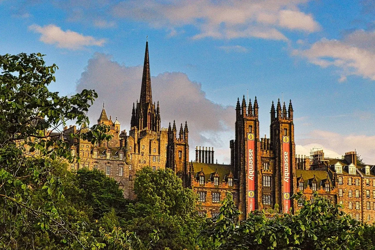Historic stone buildings and a tall, pointed spire rise above green trees in Edinburgh. Red banners with 'assembly' text h...