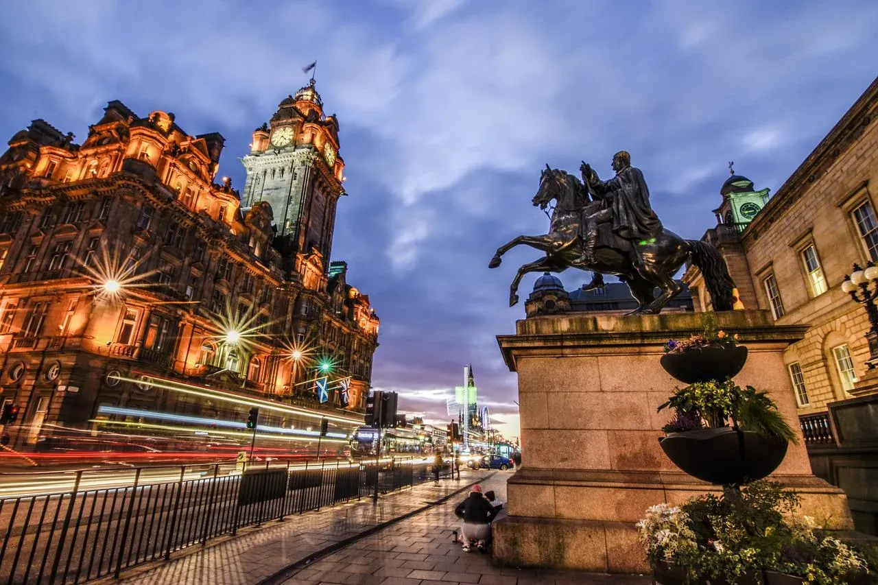 Balmoral Hotel clock tower and facade lit at dusk, with an equestrian statue, street lights, and blurred car lights on a w...