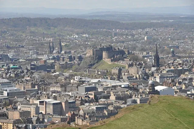 Panoramic view of Edinburgh from a hill, showing Edinburgh Castle on a rock, city buildings, and distant green hills.