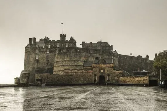 Edinburgh Castle's imposing stone facade and battlements under an overcast sky. The wet esplanade in the foreground reflec...