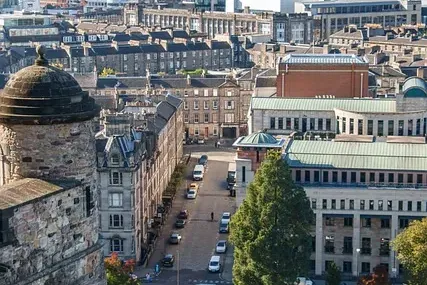 View of Edinburgh cityscape from a high vantage point, showing a stone castle tower on the left, a street with cars, and d...